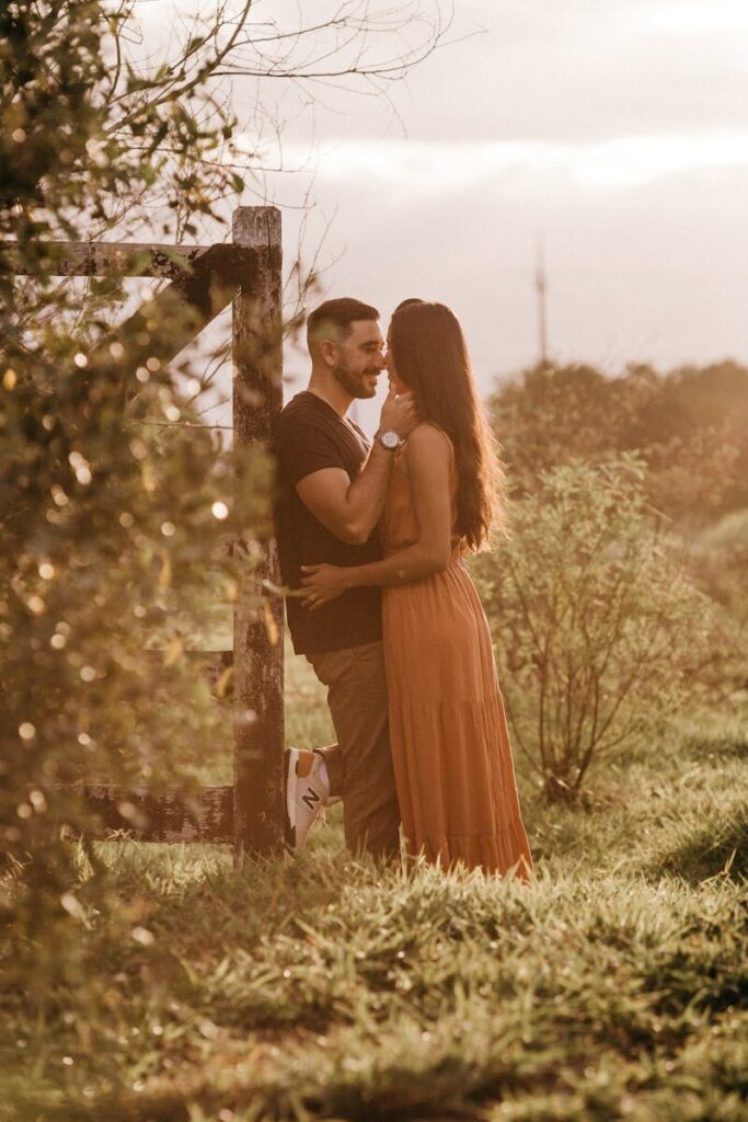 Side view of young content ethnic man caressing cheek of elegant girlfriend while looking at each other on meadow in back lit